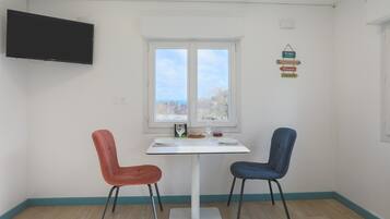 Quarto com mesa branca, duas cadeiras, televisão e janela com vista.