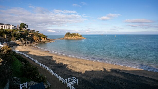 A coastal hotel with a beach view, a white fence, and a rocky outcrop in the distance.