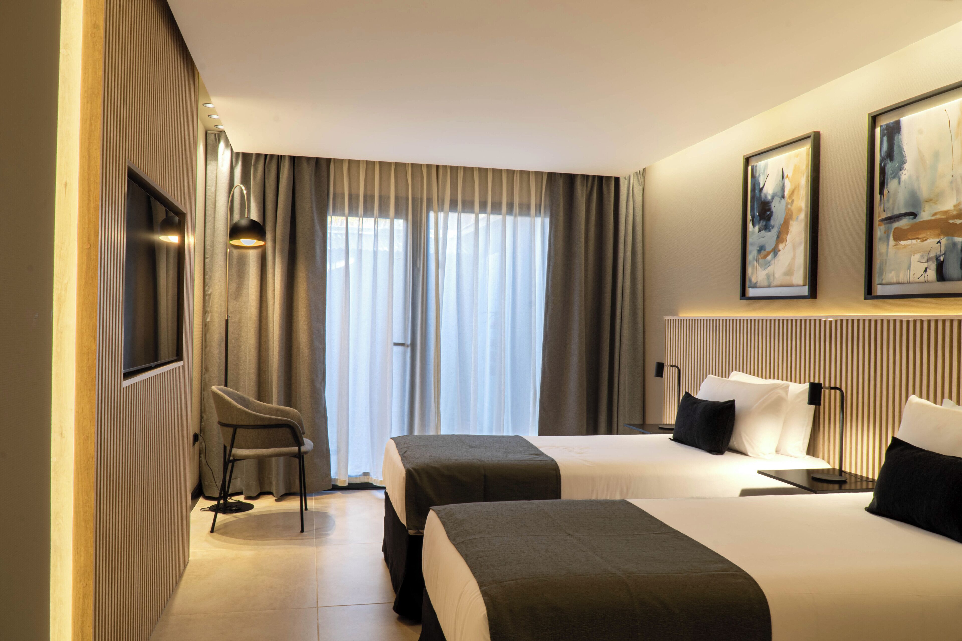 A hotel room with two beds, a chair, a TV, and artwork on the wall.