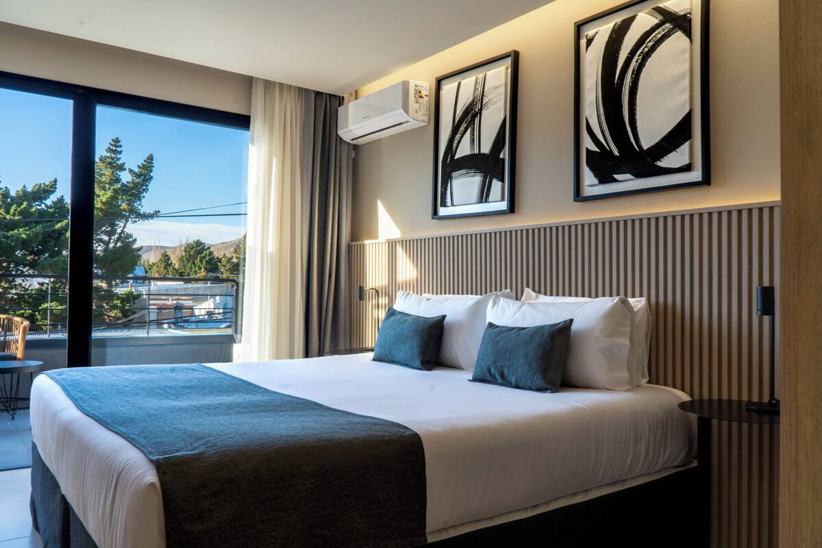 A hotel room with a large bed, two pillows, a bedside table, a lamp, and a view of trees and mountains through a large window.