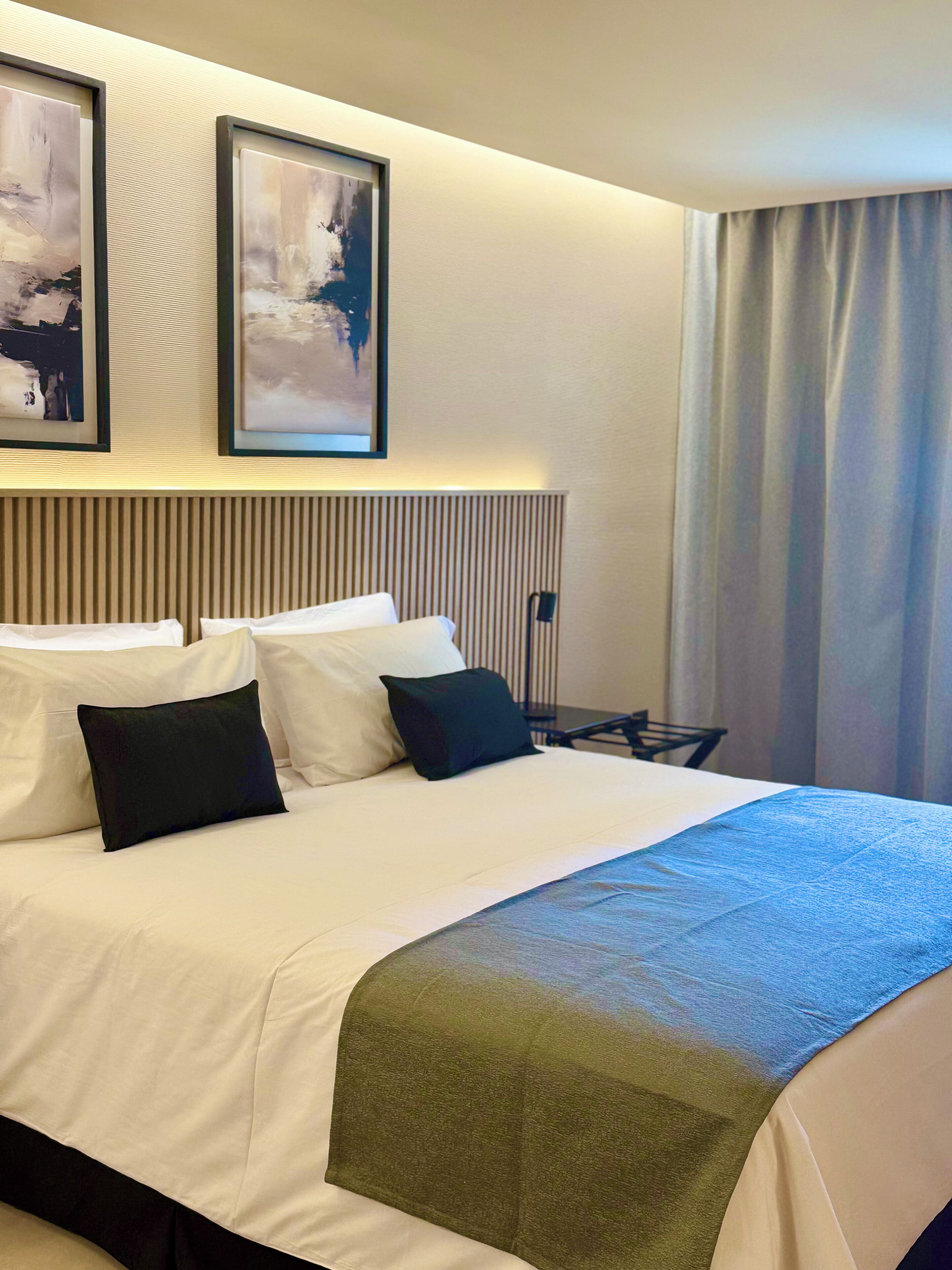 Habitación de hotel con una cama grande, dos cuadros abstractos enmarcados, un cabecero con líneas verticales y una ventana con cortinas.