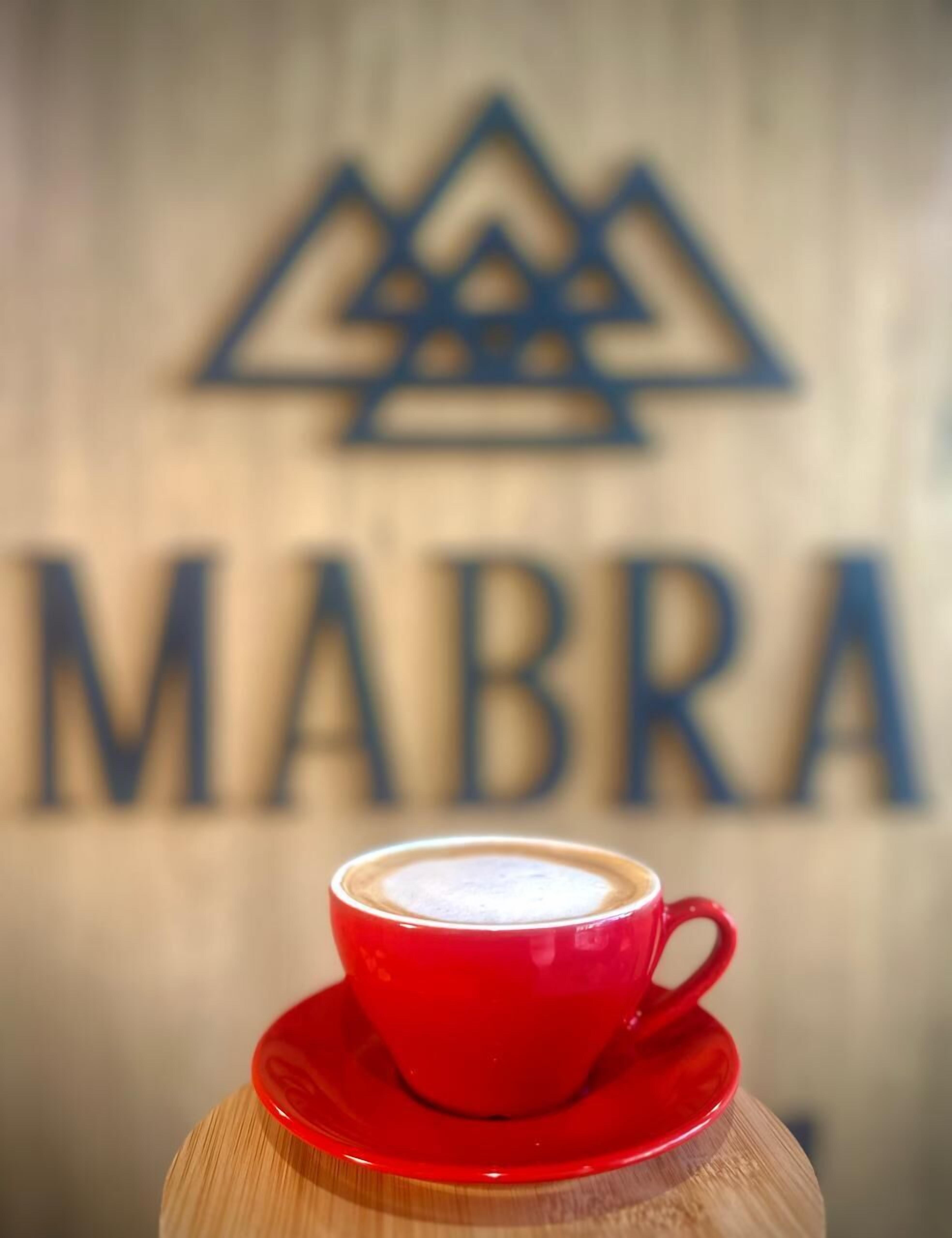 A red cup of coffee on a saucer in front of a sign with the word "MABRA".