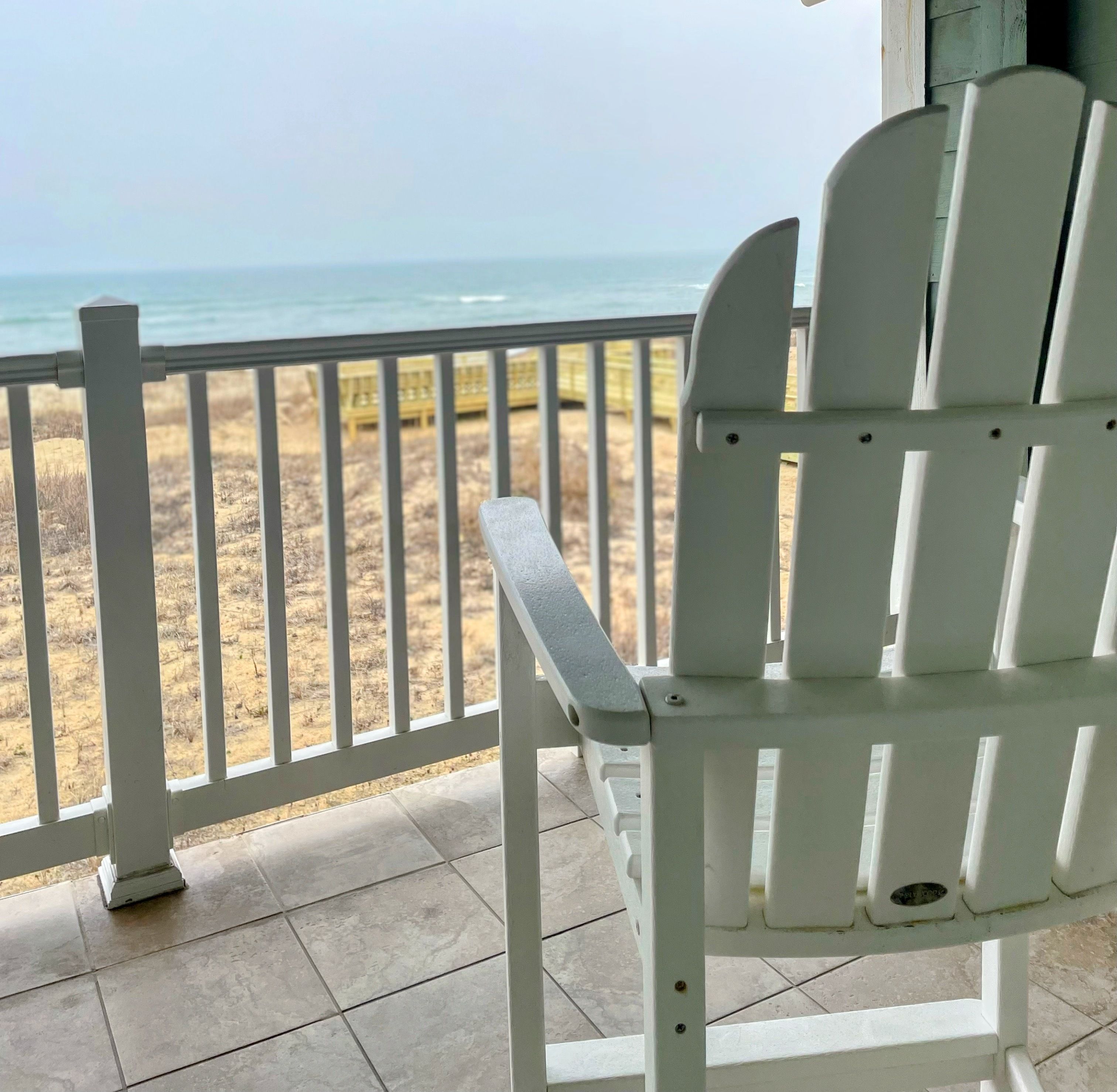 A white Adirondack chair on a tiled porch overlooking a beach with a railing.