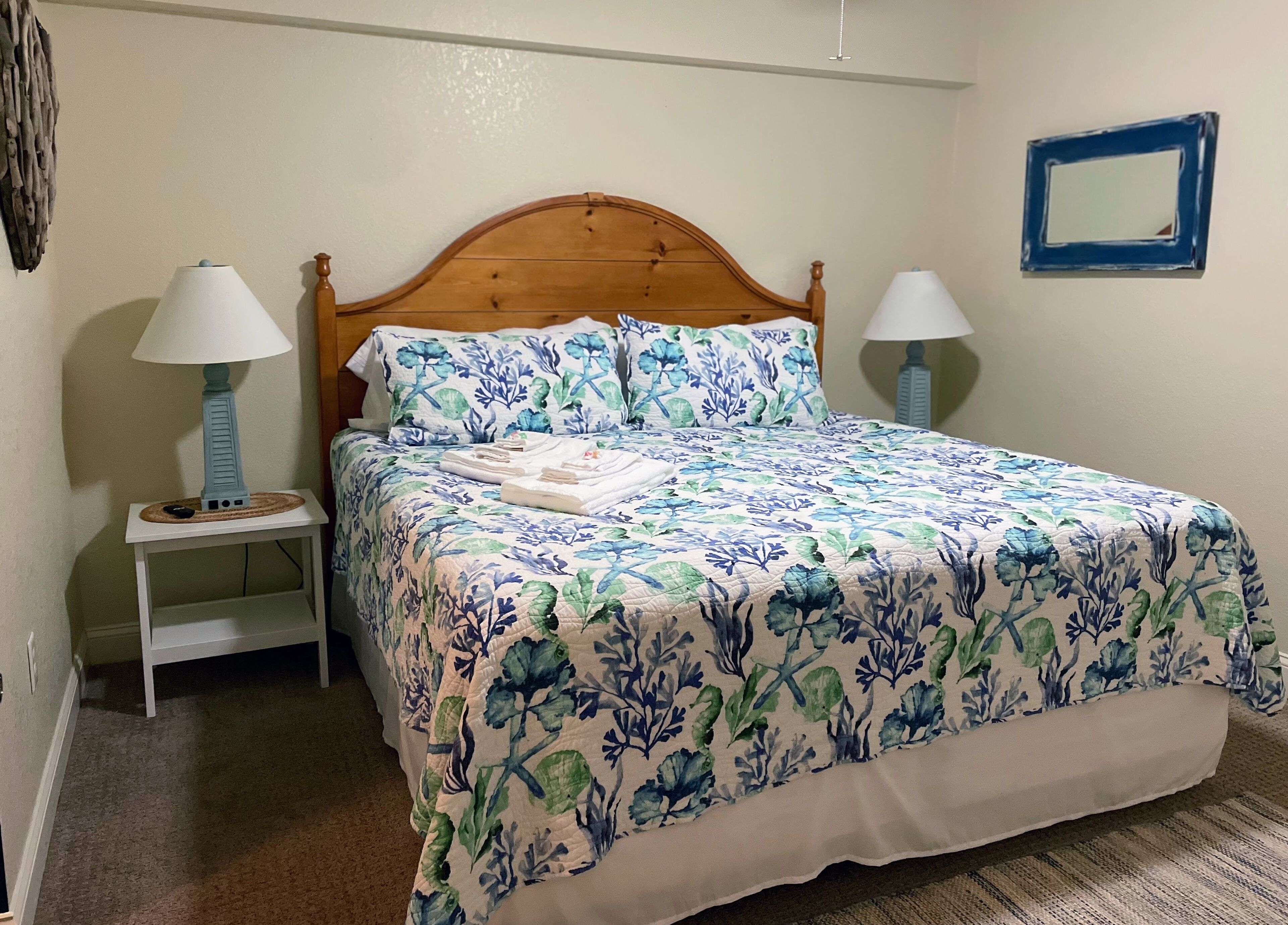 A neatly made bed with floral bedding, two bedside lamps, a small bedside table, and a wall-mounted mirror.