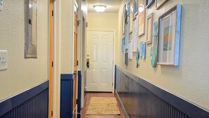 A hallway with wooden flooring, beige walls, and blue trim. There are two doors, a wall-mounted light fixture, and framed artwork on the walls. - Oceanfront paradise! Family-friendly condo with perfect unobstructed beach views (Kill Devil Hills)