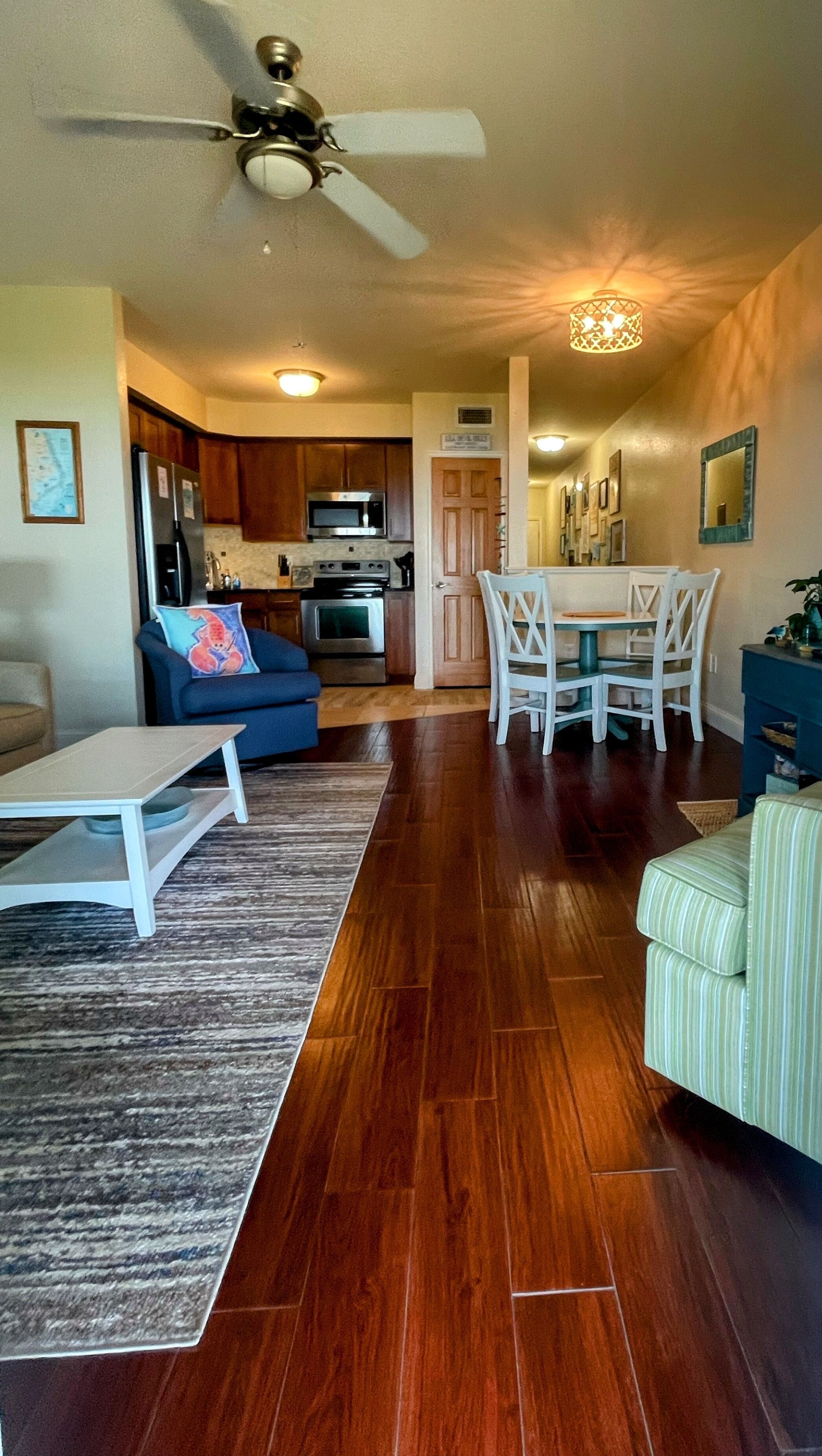 A spacious living room with hardwood flooring, a ceiling fan, a dining area with a white table and chairs, and a kitchen with wooden cabinets and appliances.