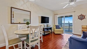 A living room with a dining table, chairs, a TV, a sofa, and a balcony with a view. - Oceanfront paradise! Family-friendly condo with perfect unobstructed beach views (Kill Devil Hills)