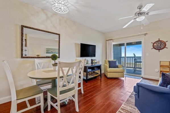 A living room with a dining table, chairs, a TV, a sofa, and a balcony with a view.
