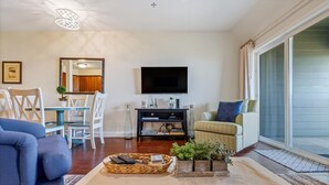 A living room with a television, a dining table with chairs, a sofa, and a large window with curtains. - Oceanfront paradise! Family-friendly condo with perfect unobstructed beach views (Kill Devil Hills)