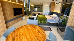 A modern hotel room with a wooden accent wall, a flat-screen TV, a sofa, a bed with a green blanket, and a dining area with a wooden table. - New and elegant studio in Campo Grande/MS (Centro Campo Grande)