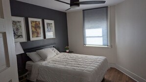 Un dormitorio con cama, ventilador de techo, ventana con persianas y tres cuadros en la pared.