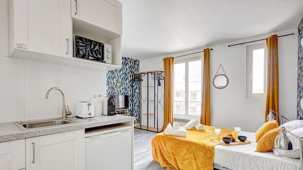 A compact hotel room with a kitchenette, a bed with a yellow blanket, a window with curtains, and a view of the city. - TBS 142 - Cosy Studio at the Gates of Paris (Paris)