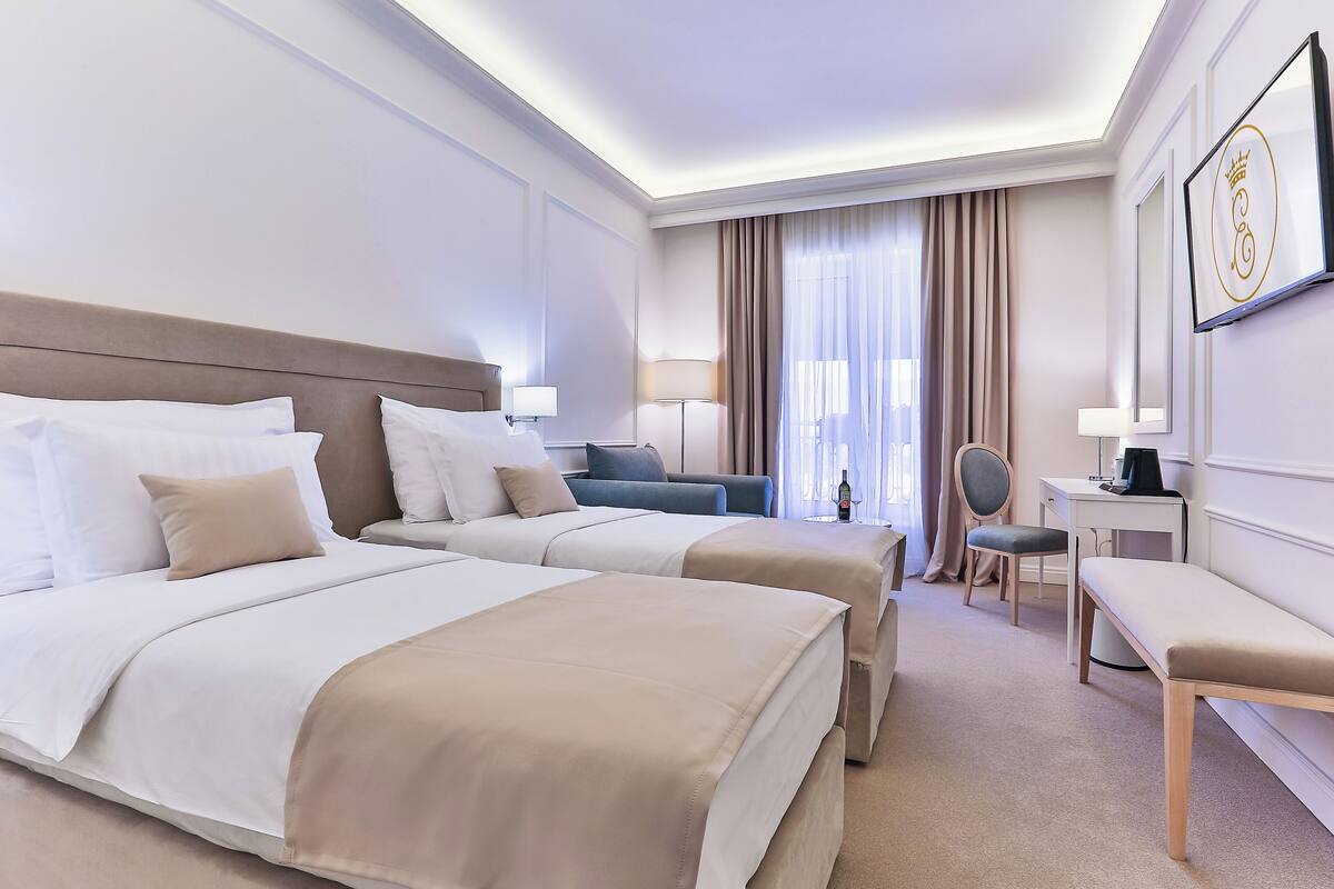 Ein Hotelzimmer mit zwei Betten, einem Schreibtisch, einem Sessel und einem großen Fenster mit Vorhängen.