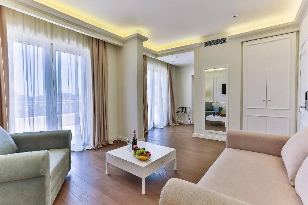 Ein geräumiges Wohnzimmer mit einem weißen Couchtisch, beigefarbenen Sofas und großen Fenstern mit transparenten Vorhängen.