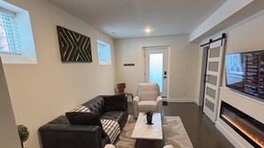 A modern living room with a black leather sofa, a white armchair, and a coffee table. There is a fireplace on the right and a large TV screen on the wall. - Newly-built modern farmhouse condo available in Grosse Pointe Park! (Grosse Pointe Park)