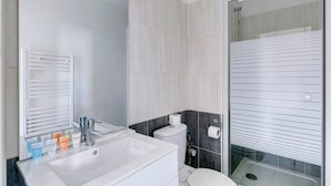 A modern bathroom with a white sink, mirror, and toilet, and a glass-enclosed shower area. - TheBlueStay 135 - Cosy apartment (Paris)
