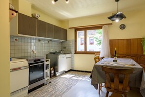 Una cocina con estufa, horno y lavavajillas, una mesa de comedor con sillas, una ventana con cortinas y un reloj en la pared.