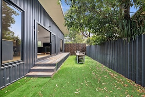 A modern house with a deck, a dining area, and a fenced backyard.