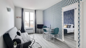 A modern hotel room with a sofa, a dining table with chairs, a TV, and a bed with a patterned headboard.