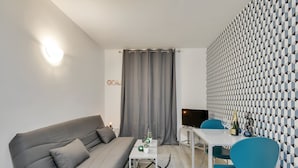 A modern living room with a grey sofa, a white coffee table, and a TV on a stand. There is a patterned wall and a curtain in the background.