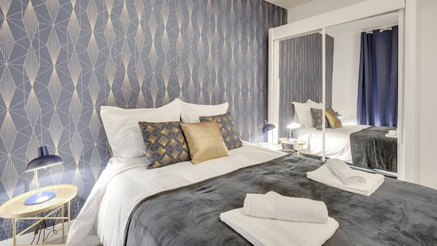 A hotel room with a geometric patterned wallpaper, a bed with white bedding and decorative pillows, a bedside table with a lamp, and a large mirror reflecting the room.