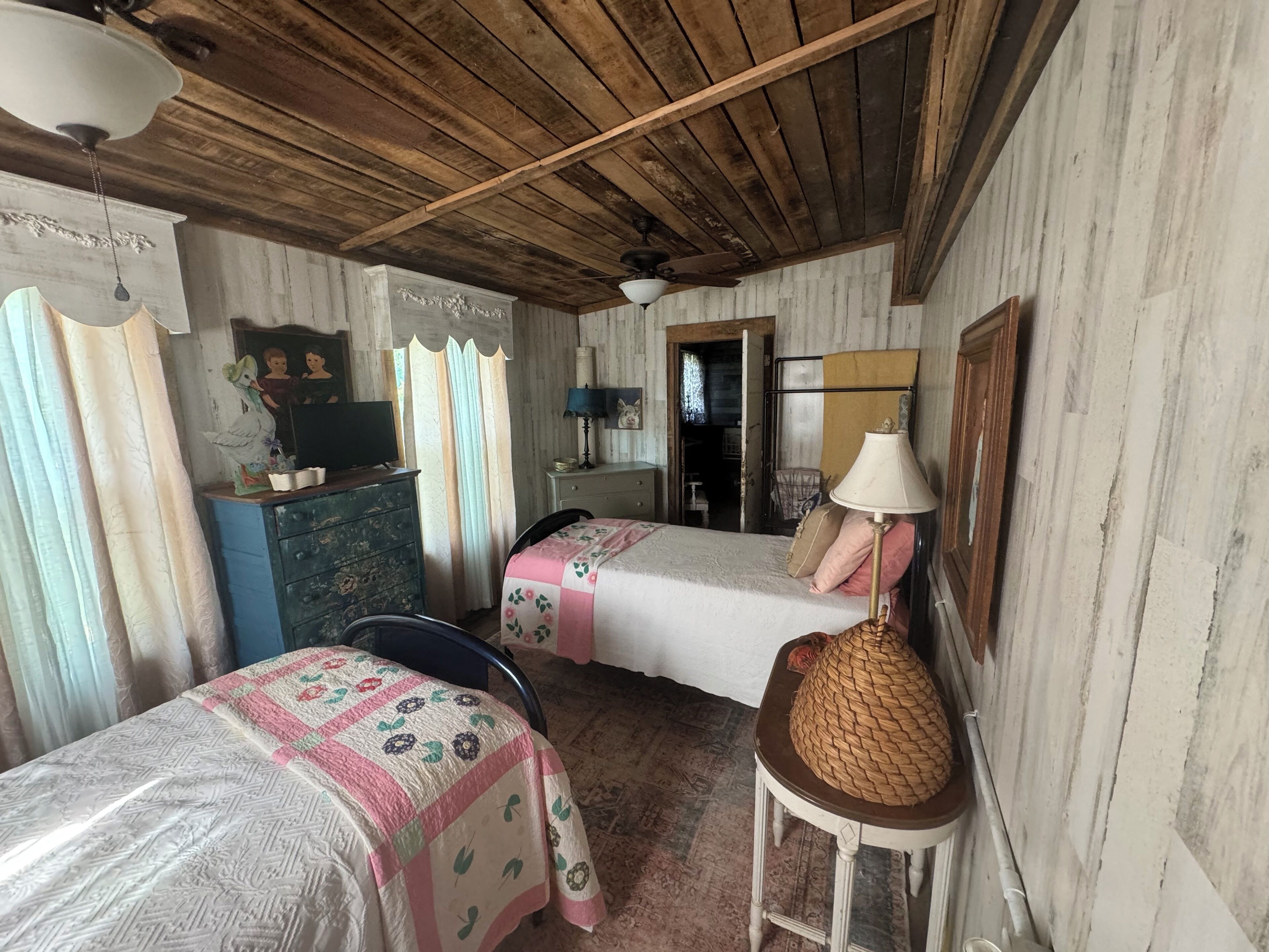 Una habitación rústica con vigas de madera en el techo, dos camas con ropa de cama floral, un tocador, una lámpara y una cesta tejida.