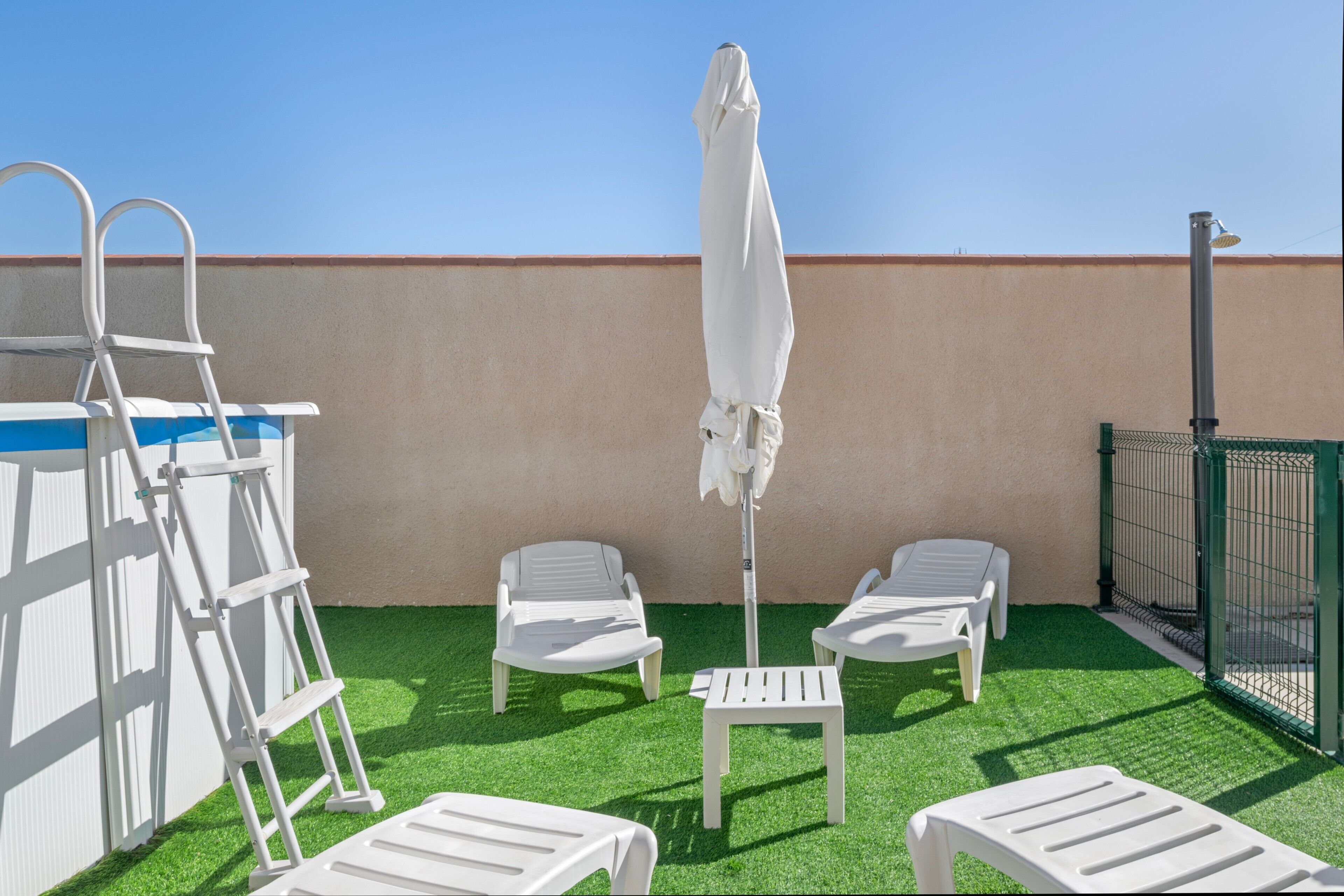 Une terrasse sur le toit, avec un revêtement d’herbe artificielle, des fauteuils blancs, un parasol blanc et une échelle en métal.