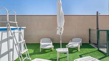 Une terrasse sur le toit, avec un revêtement d’herbe artificielle, des fauteuils blancs, un parasol blanc et une échelle en métal.