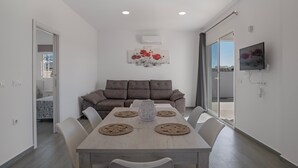 A modern living room with a sofa, a dining table with placemats, a TV mounted on the wall, and a sliding glass door leading to a balcony.