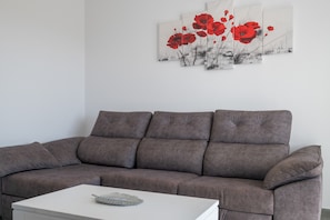 Een grijze bank met een witte salontafel ervoor, een muur met een schilderij van een klaproos, en een witte muur.