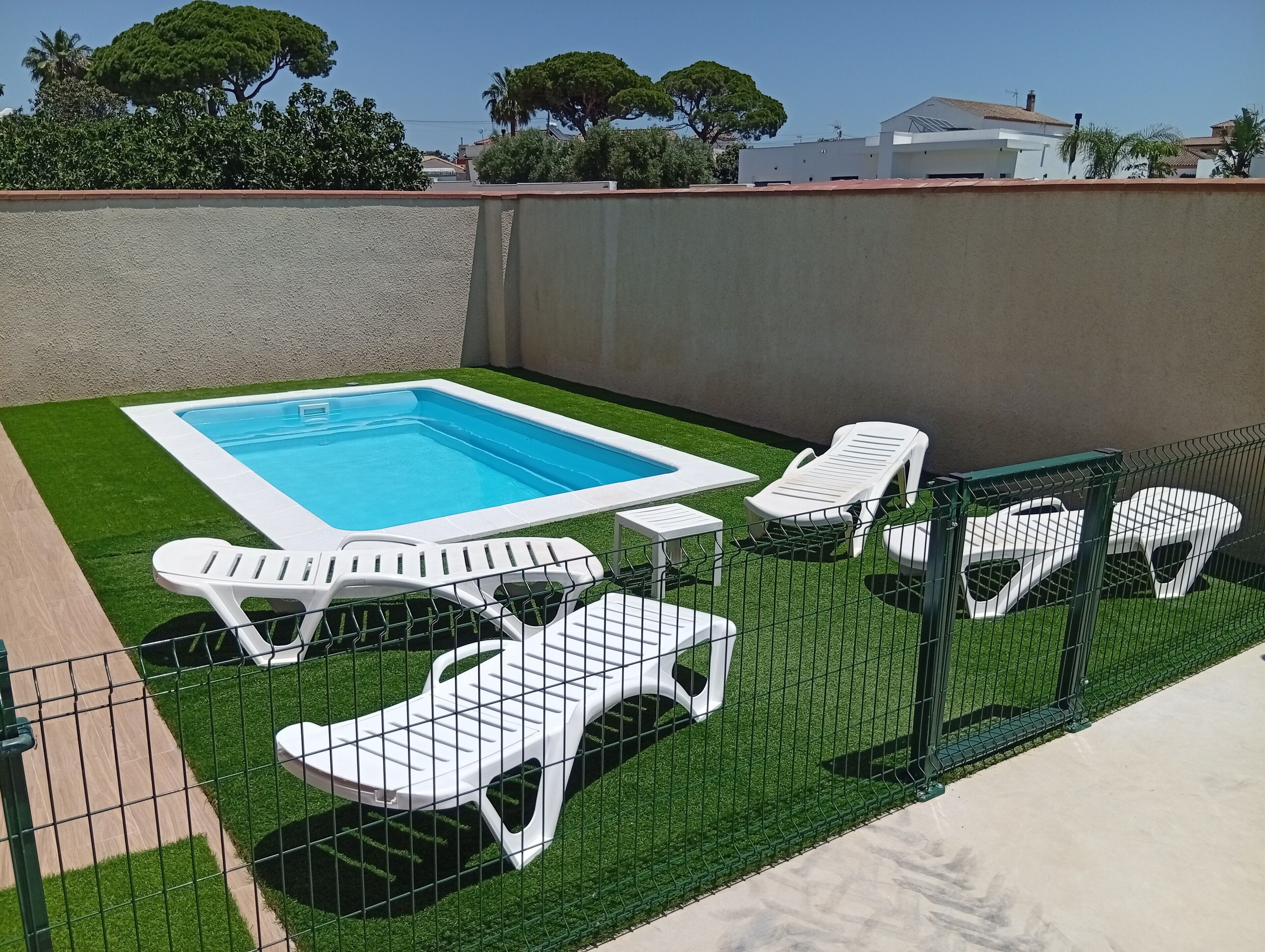 En swimmingpool med lille terrasse, omgivet af et grønt hegn, samt hvide liggestole.