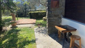 Un bâtiment en pierre avec une table et des chaises en bois à l’extérieur, un pot de fleurs et un chemin de briques menant à une haie.