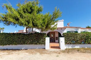 A white building with a tiled roof, a large tree in front, and a hedge along the side.