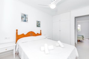 A bedroom with a wooden headboard, white bedding, a bedside table, a ceiling fan, and a wardrobe.
