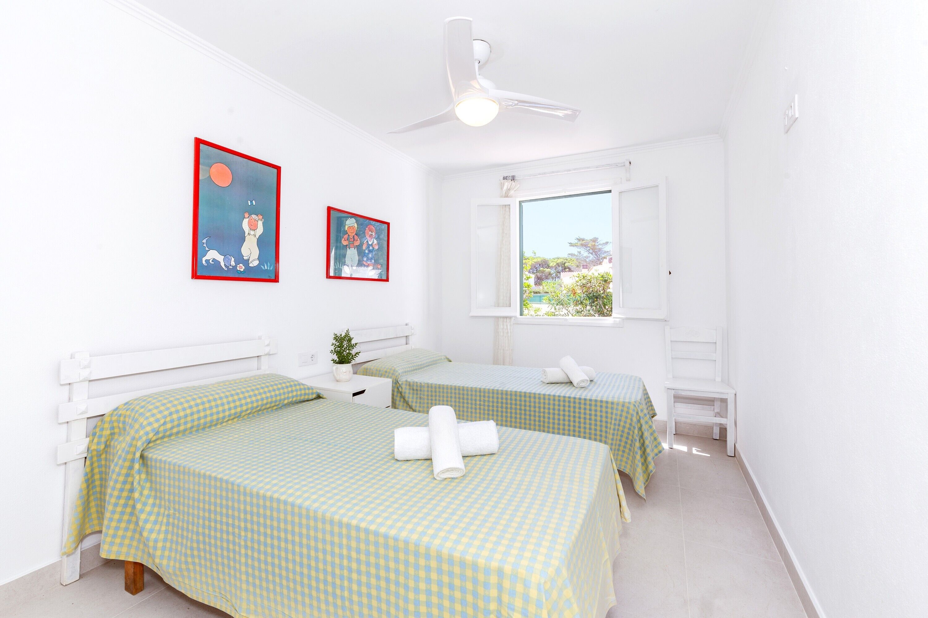 Una habitación con dos camas, un ventilador de techo, una ventana con vistas y dos cuadros en la pared.