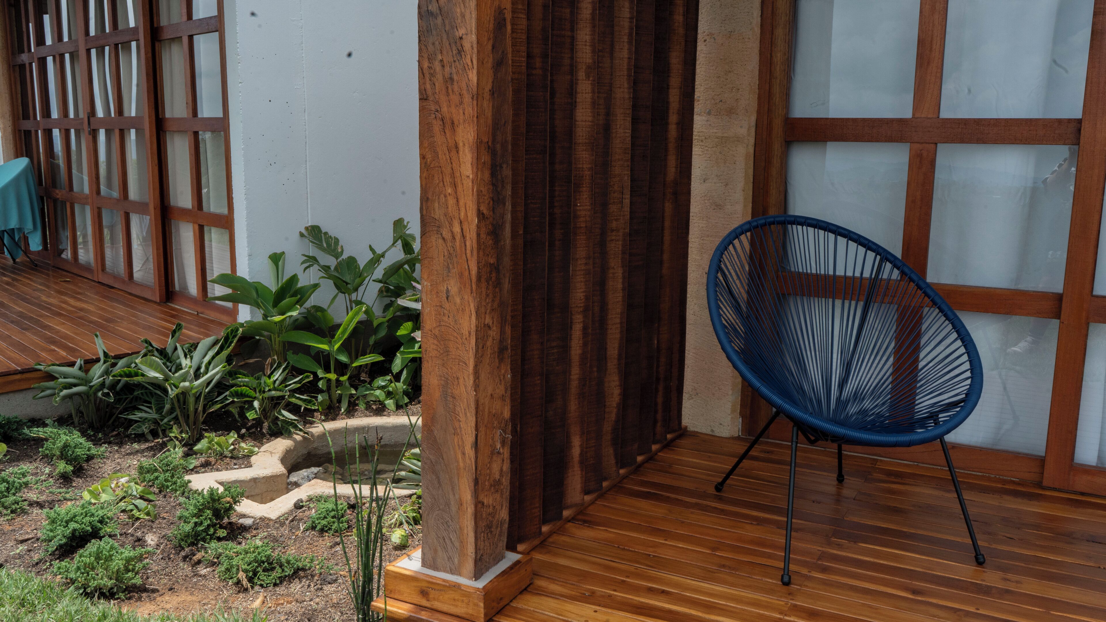 Una terraza de madera con una silla azul, un pequeño jardín con diversas plantas y una cerca de madera.