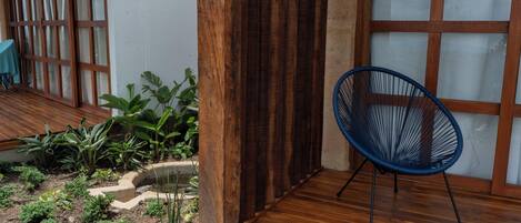 Una terraza de madera con una silla azul, un pequeño jardín con diversas plantas y una cerca de madera.