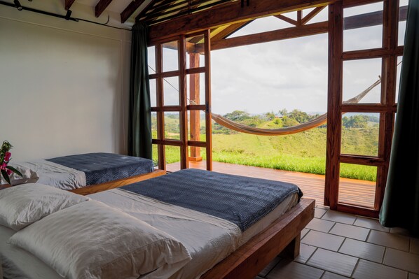 A bedroom with two beds, a hammock, and a view of a green landscape through large windows. - Hotel Cacaobetulia (Maceo)