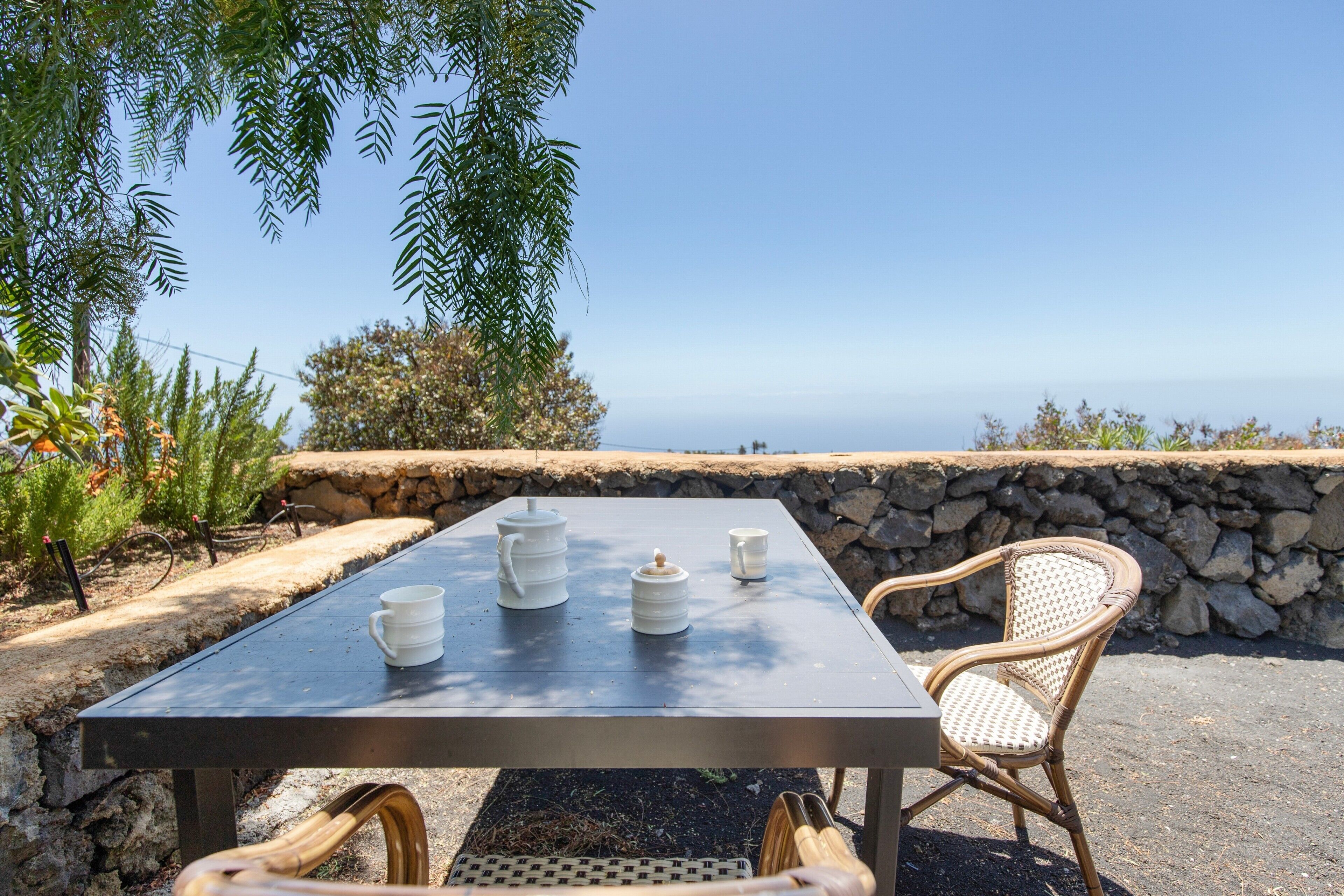 A table with a view of the ocean, a stone wall, and wicker furniture.