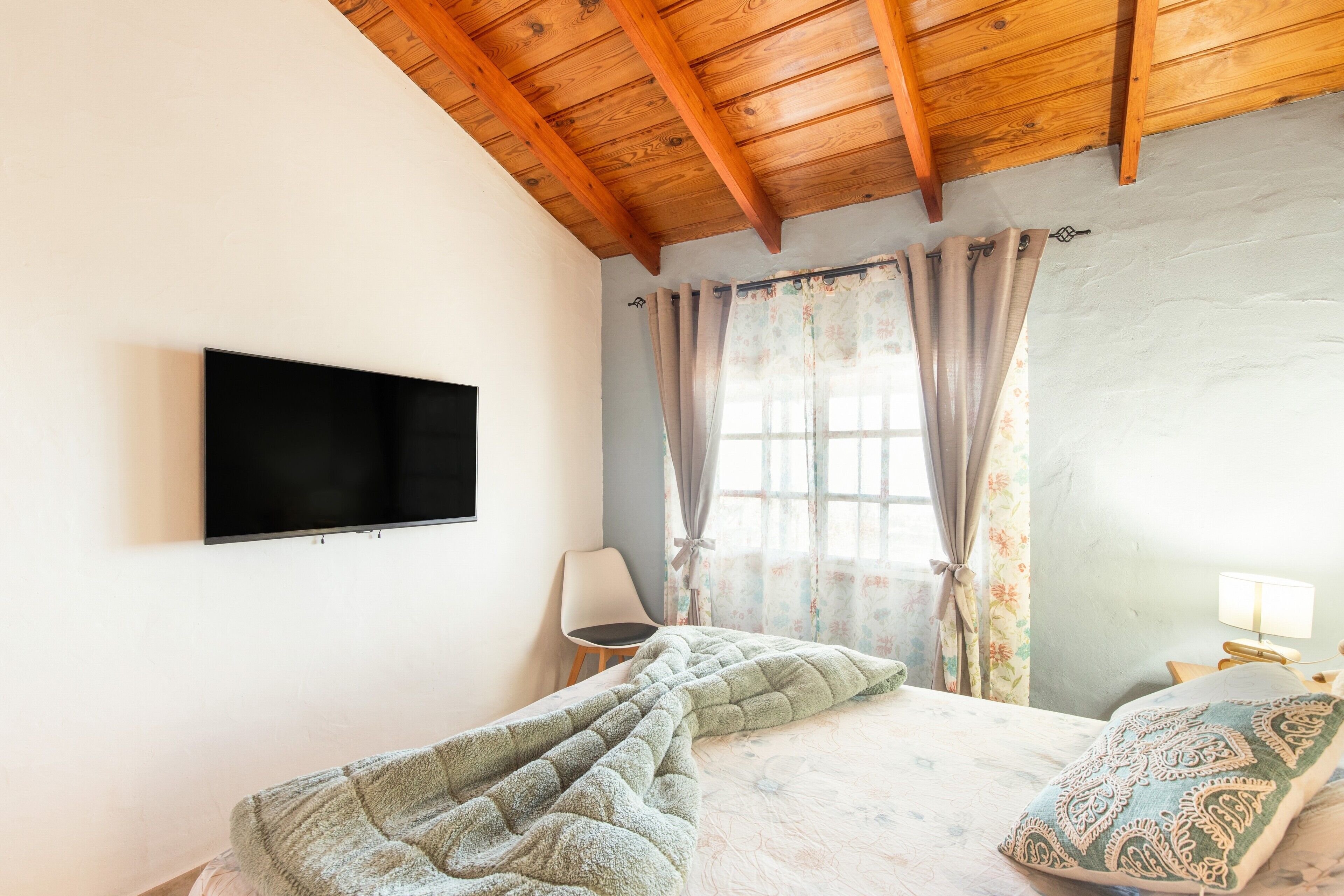 A bedroom with a bed, a chair, a TV, and a window with curtains.