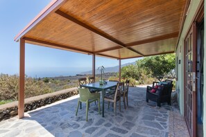 A covered outdoor seating area with stone flooring, a table, chairs, and a view of the landscape.