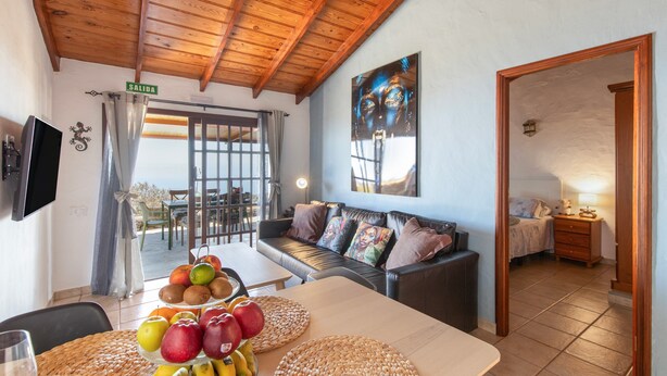 Ein Zimmer mit Holzboden, einer Couch mit Kissen, einem Esstisch mit Obst und einem an der Wand montierten Fernseher.