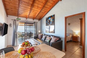 A room with a wooden ceiling, a sofa with cushions, a dining table with fruit, and a TV mounted on the wall.