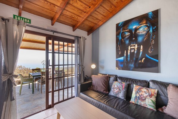A room with a wooden ceiling, a sofa with colorful cushions, a large painting on the wall, and a view through a door.