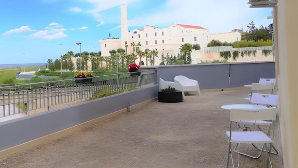 Una terraza en la azotea con sillas y mesas blancas, una barandilla y vistas a un edificio grande con techo rojo.