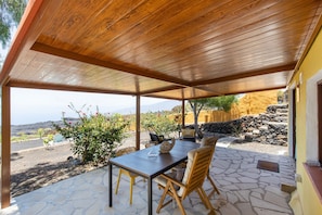 A covered patio with a table and chairs, a stone floor, and a view of the landscape.