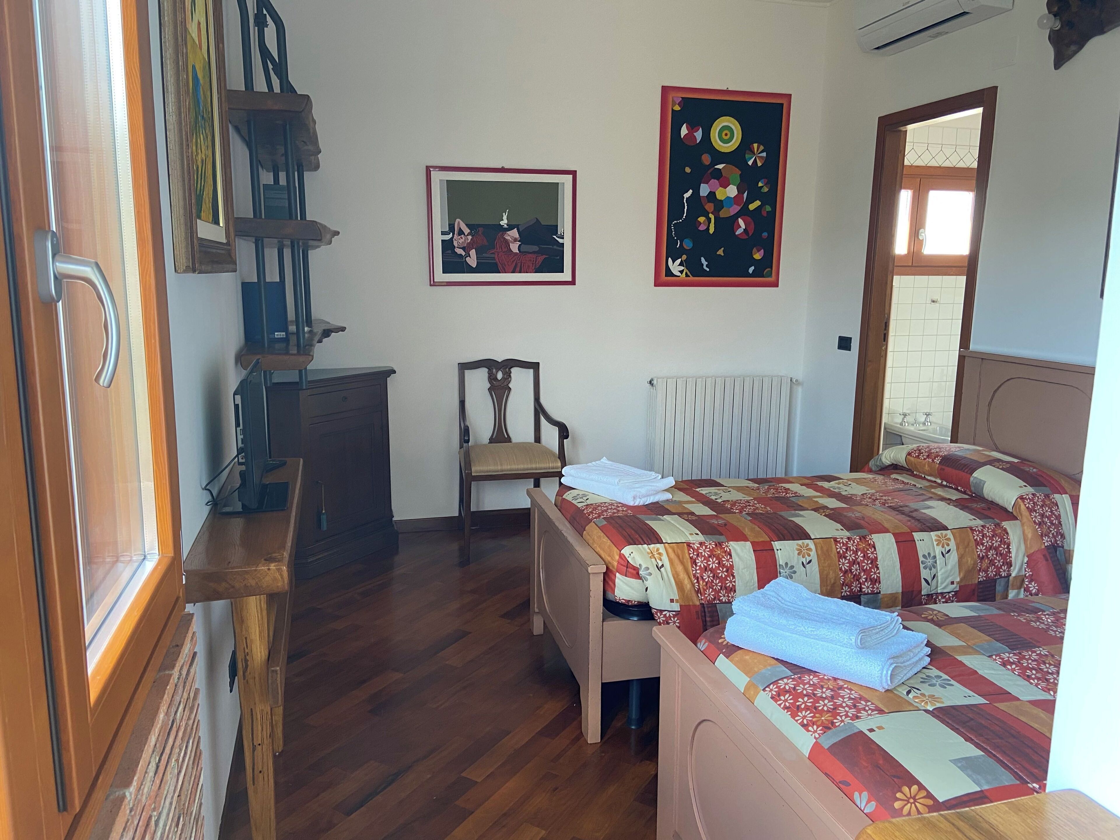 A bedroom with a wooden floor, a bed with a quilt, a chair, a radiator, and two framed artworks on the wall.