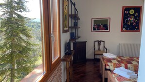 Una habitación con piso de madera, una cama con colcha, una silla, un escritorio con computadora, una ventana con vista a árboles y un cuadro en la pared.