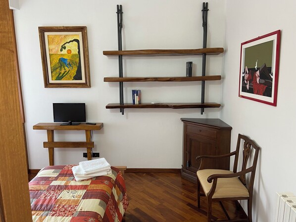 Un dormitorio con una cama, una silla, un escritorio con televisor y estanterías con libros.