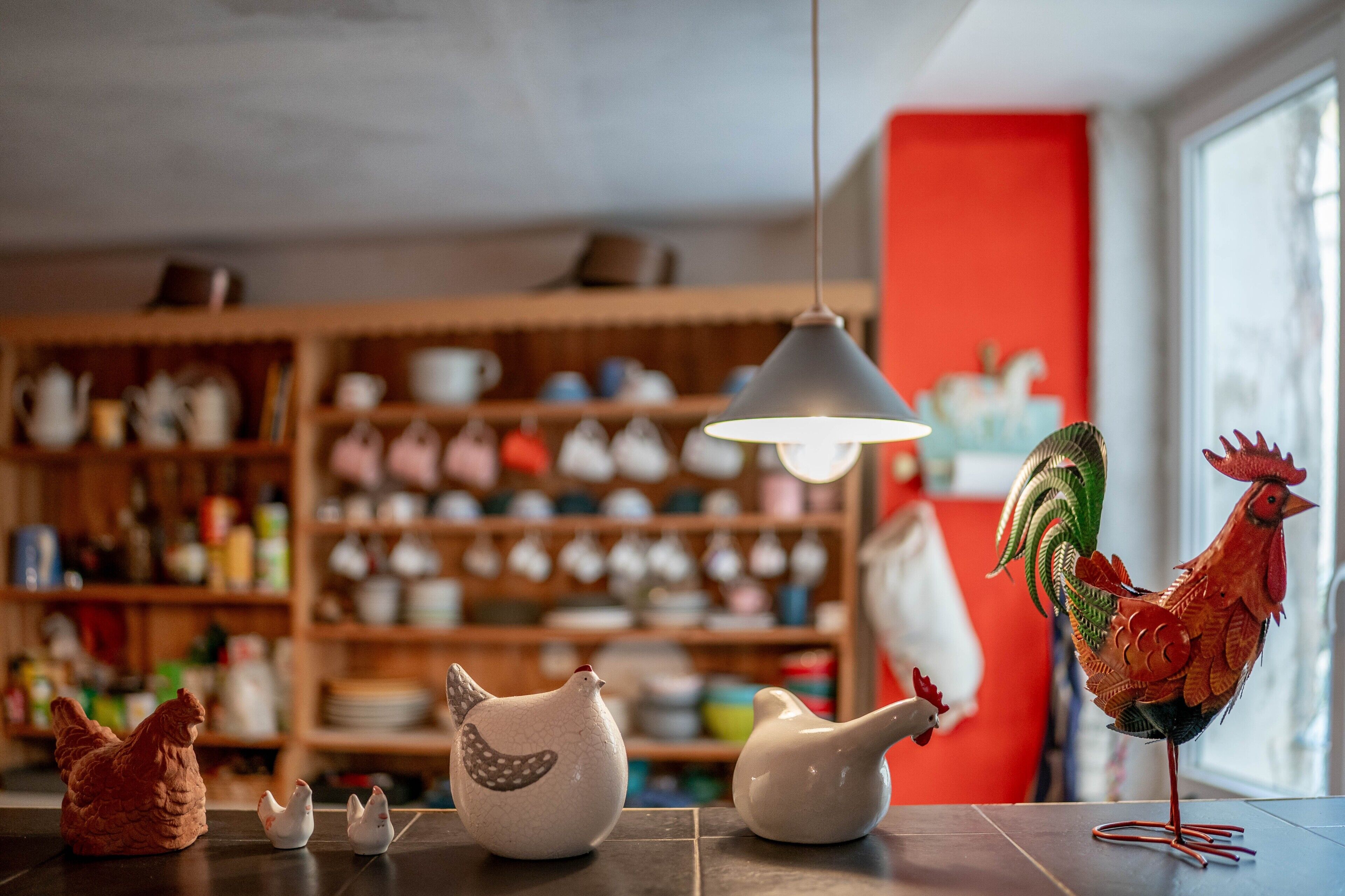 Une cuisine décorée de statuettes en céramique représentant des poulets et des coqs, d’un luminaire suspendu et d’étagères remplies d’ustensiles de cuisine.